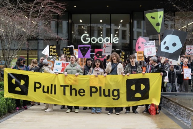 A photo of a large group of people posing together in front of a black glass building with the name Google on it in white. The people hold protest signs and a yellow banner that reads "Pull the Plug".