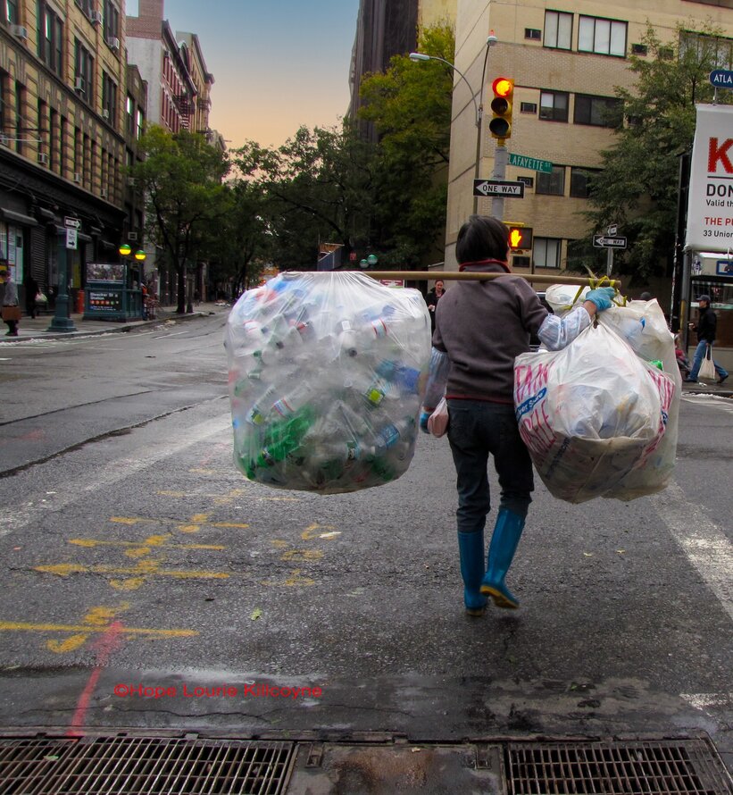 Uma pessoa atravessa a rua na cidade de Nova Yorque ao anoitecer. Elescarregam uma vara de madeira no ombro direito, com dois enormes sacosplásticos brancos translúcidos equilibrados em cada extremidade, repletos degarrafas plásticasvazias.