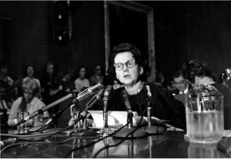 A black and white photo of Rachel Carson in middle age sitting at a table and speaking into a group of microphones on small stands. Papers sit before her, probably notes. There is a pitcher of water nearby. There is a crowd of people sitting or standing behind her. The details of the room are dim and hazy.