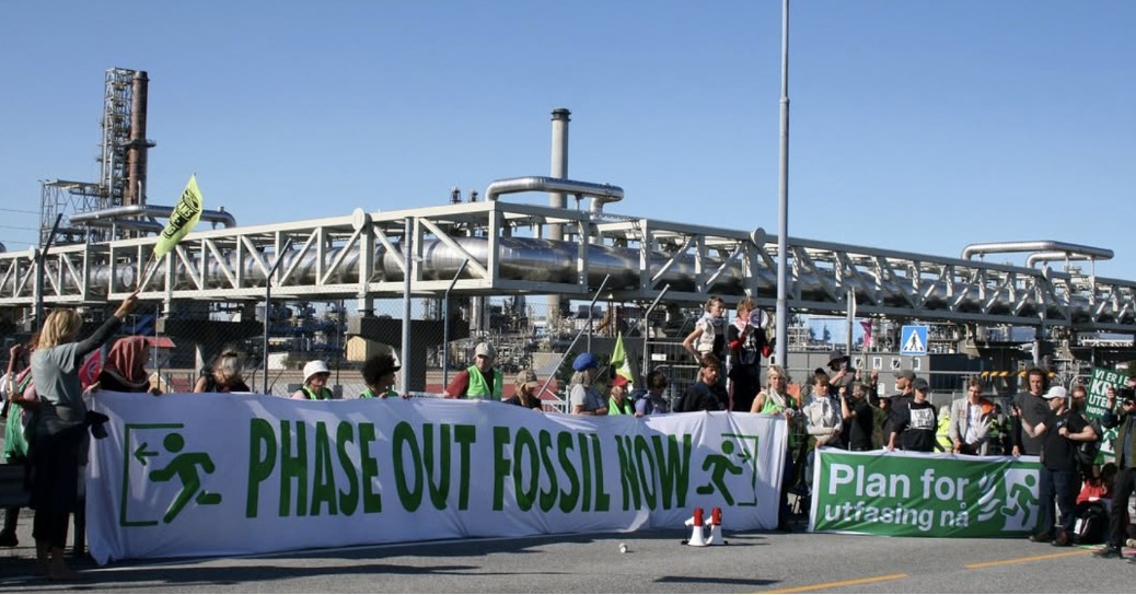 A photograph of a group of people standing in front of some kind of industrial complex dominated by both horizontal and vertical pipes. The people hold green and white banners, one of which reads "phase out fossil now." The other is too wrinkled to read in its entirety, but it includes the words "plan for." Both feature a symbol suggesting a human running through a door. Some also hold smaller signs that can't be clearly read. Two megaphones sit on the ground. The sky is clear blue.