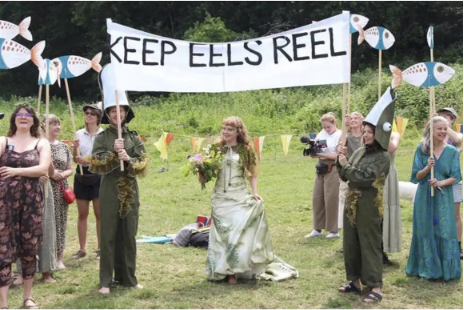A photo of what looks vaguely like an outdoor wedding, though only a single bride is visible and no groom. The people standing on either side of the bride are wearing costumes that are probably meant to represent eels as they hold a banner above the bride that reads "keep eels reel." They don't look very eel-like, though. A few other people stand around holding large, paper fish on poles. One person holds a video camera. Everything looks very festive and funky.